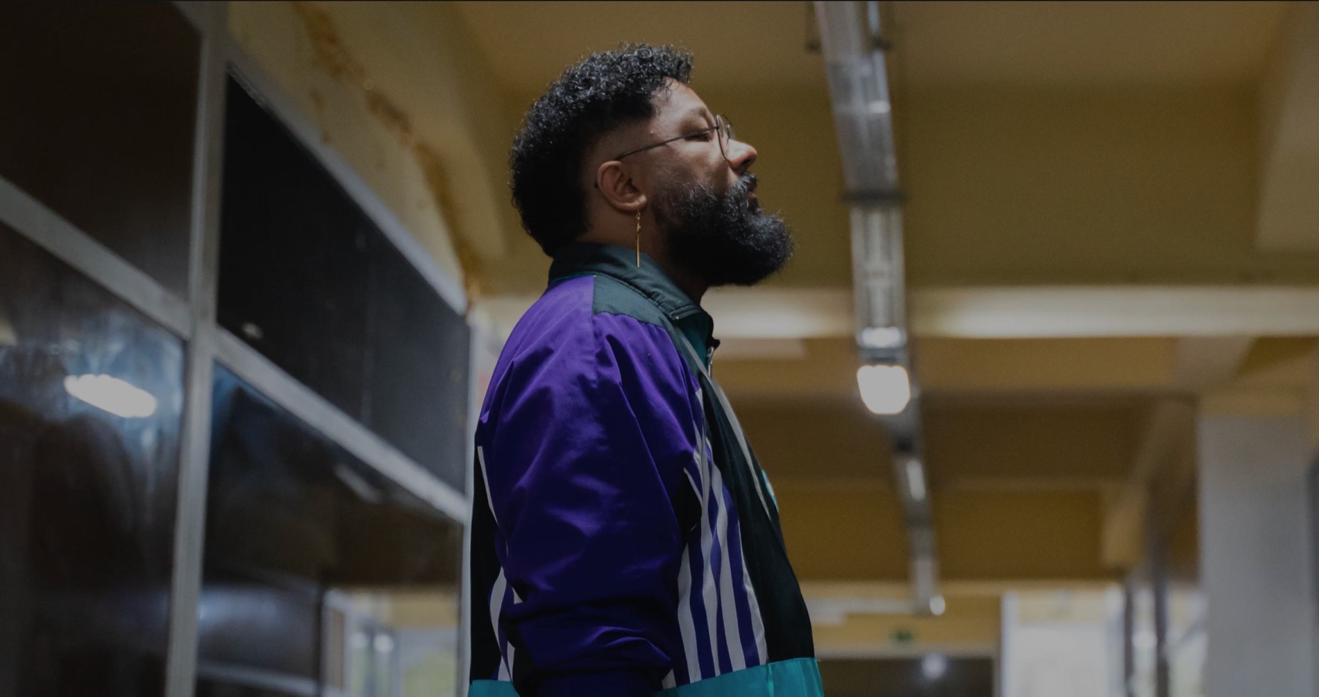 Azandr de profil dans un couloir industriel, veste de survêtement violette et verte.