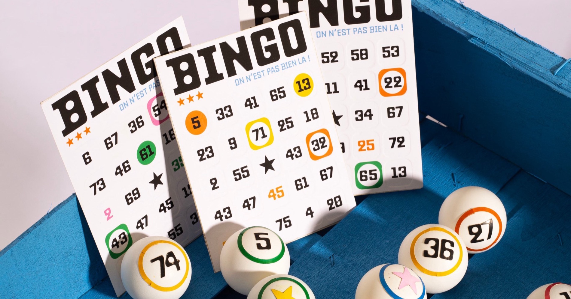 Trois cartons de Bingo dressés dans une caisse en bois bleue, entourés de boules de loto.