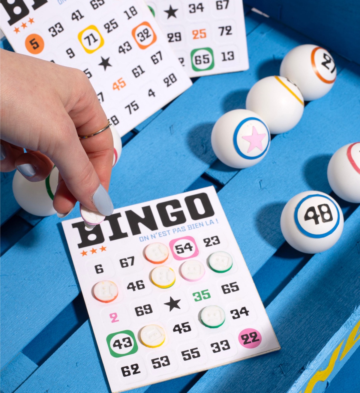 Une main pose un jeton sur un carton de Bingo, au milieu des boules éparpillées sur le banc.
