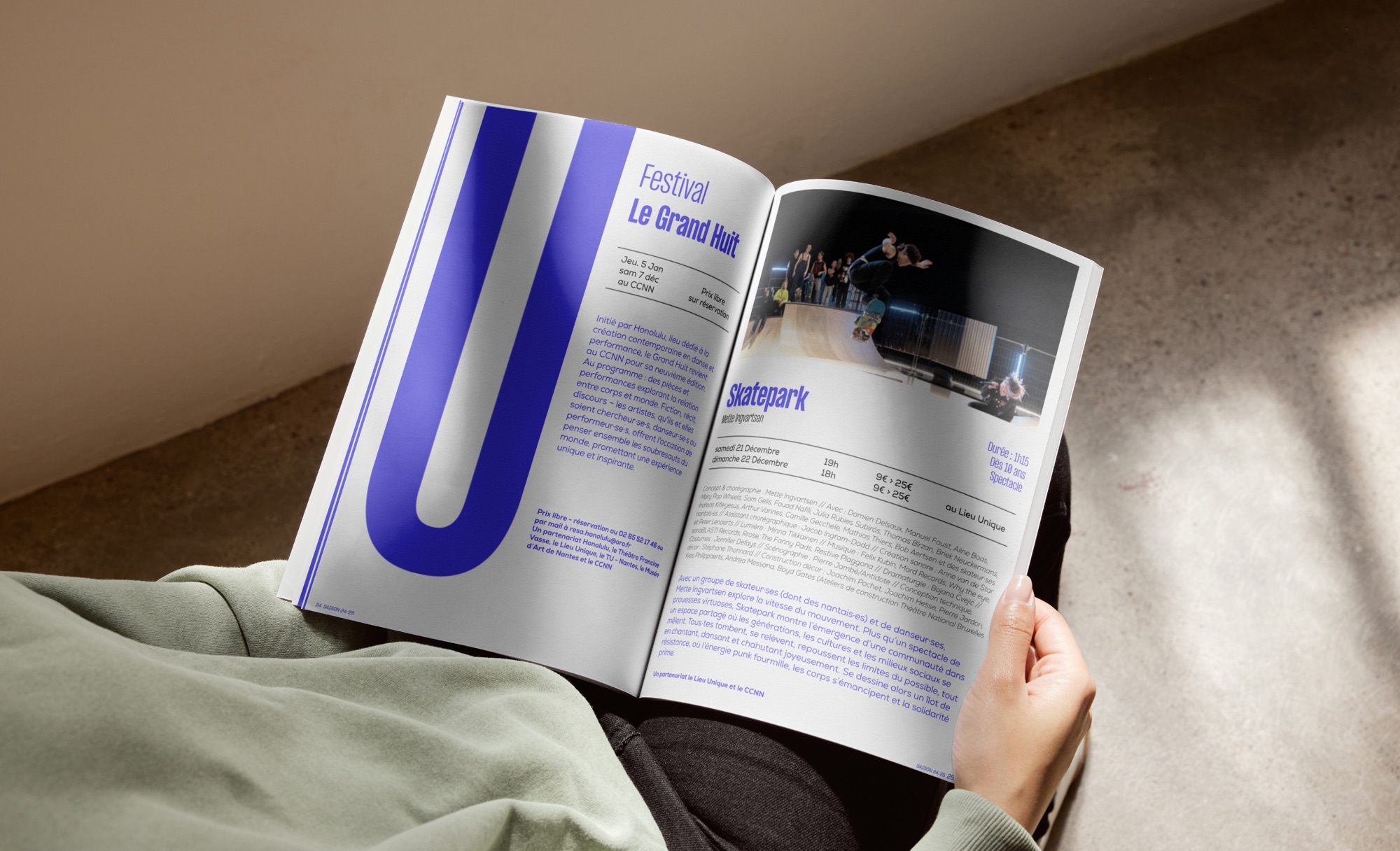 Une personne assise au sol feuillette le programme, ouvert sur la double page du festival Le Grand Huit.