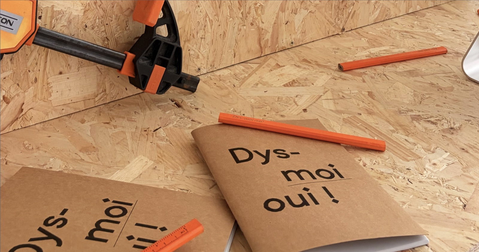 Deux livrets « Dys-moi oui ! » sous couverture kraft, posés sur un panneau d’OSB entre un serre-joint et des crayons de charpentier.