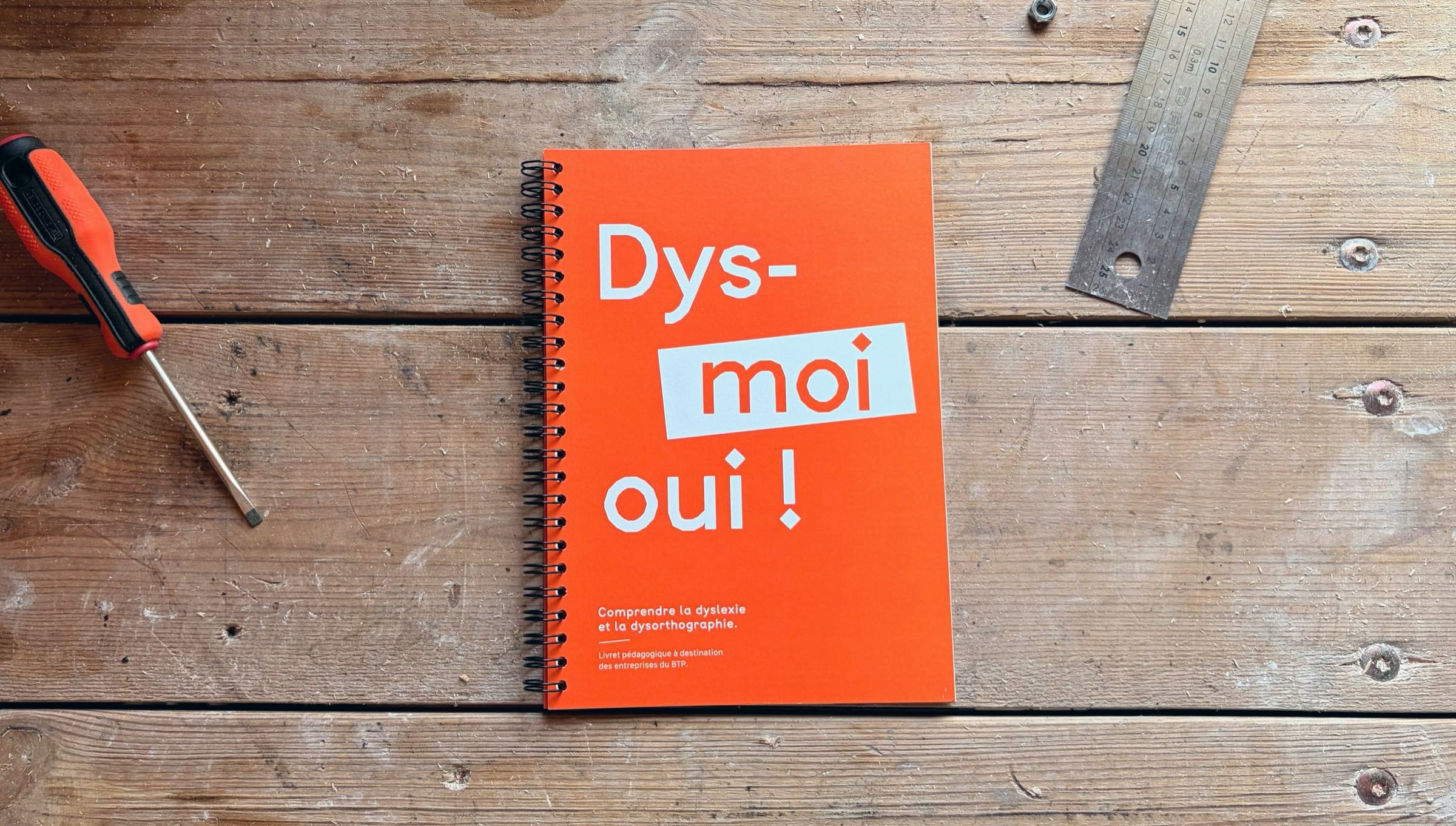 Le livret « Dys-moi oui ! », couverture orange à reliure spirale, posé sur un plateau de bois entre un tournevis et un mètre pliant.