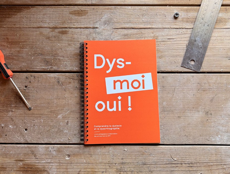 Dys-moi oui ! — carnet à spirale orange posé sur un établi en bois, entre un tournevis et un réglet.