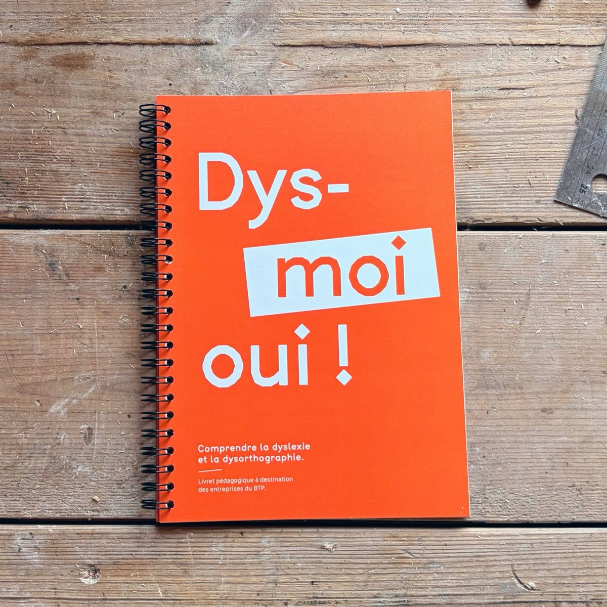 Dys-moi oui ! — carnet à spirale orange posé sur un établi en bois.