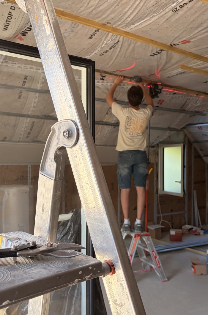 Un apprenti sur un escabeau pose une ossature au plafond d’une pièce en travaux.