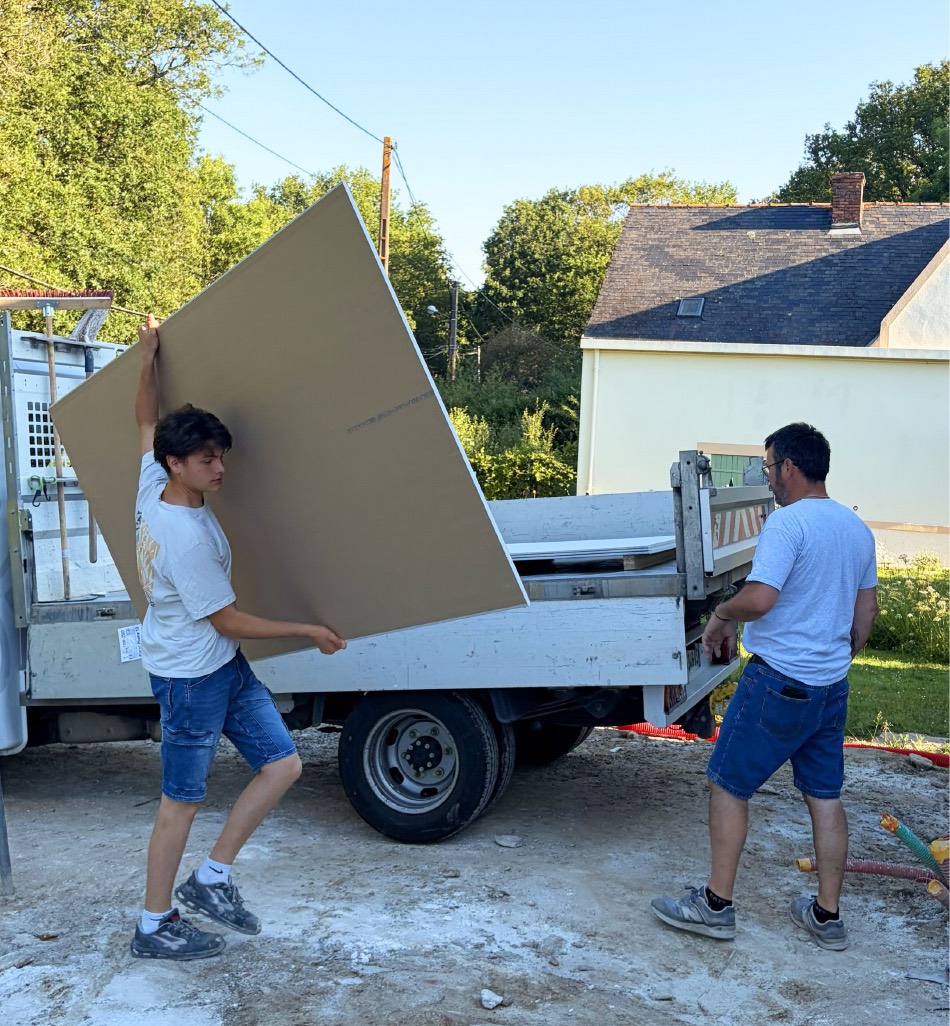 Deux hommes déchargent une plaque de plâtre depuis un camion benne, devant un pavillon.