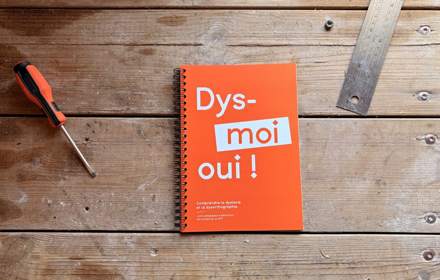 Carnet à spirale orange posé sur un établi en bois, entre un tournevis et un réglet.